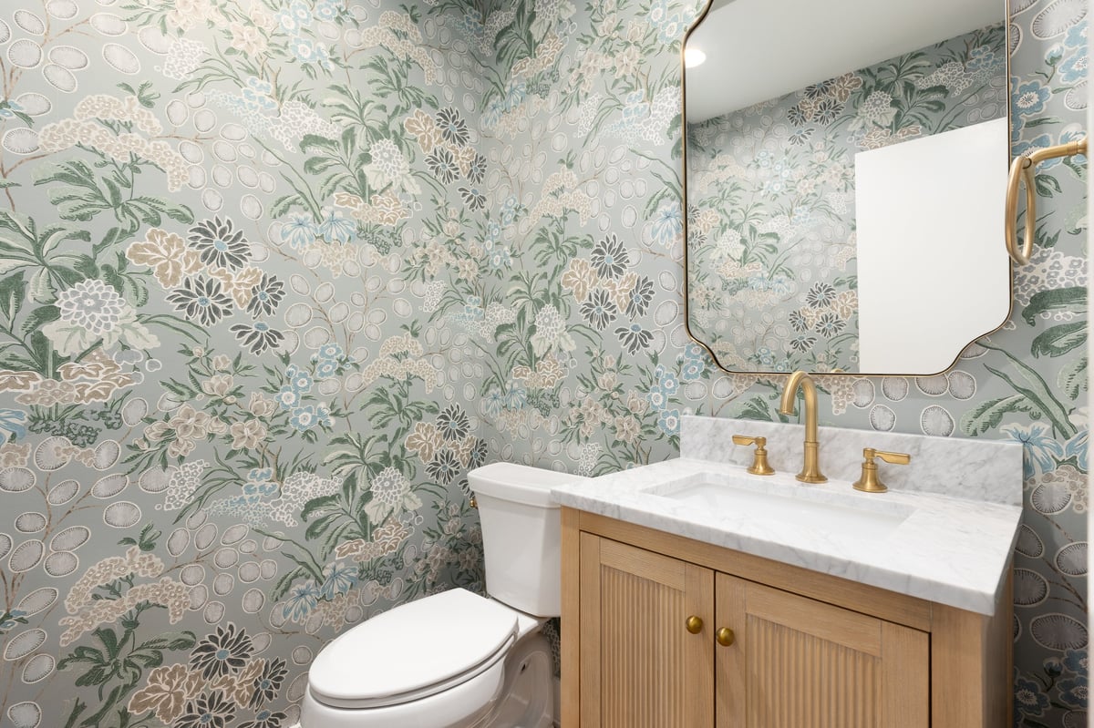 A half bathroom with a marble countertop on a white oak cabinet, gold faucet and mirror